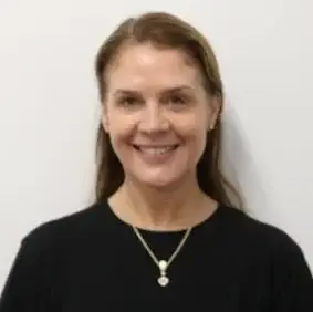 Portrait of a smiling woman with brown hair, wearing a black top and a gold pendant necklace against a light background.