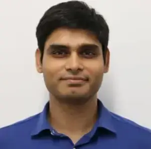 Portrait of a man with dark hair wearing a blue collared shirt against a light background.