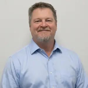 Smiling middle-aged man in a light blue button-down shirt, against a neutral gray wall.