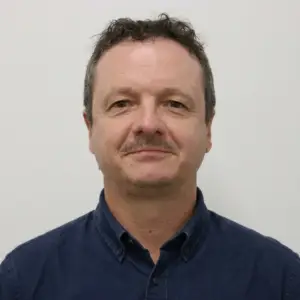 Portrait of a middle-aged man with short, dark curly hair and light facial hair, wearing a dark blue button-up shirt against a light neutral background.